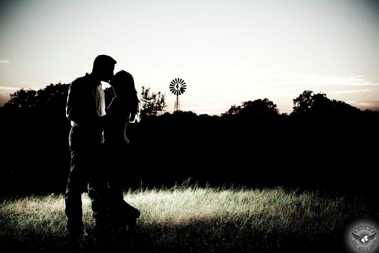 silhouette of romantic couple kissing in front of a field of wheat and a big cloudy sky with a windmill in the background in this engagement in the country of southern texas